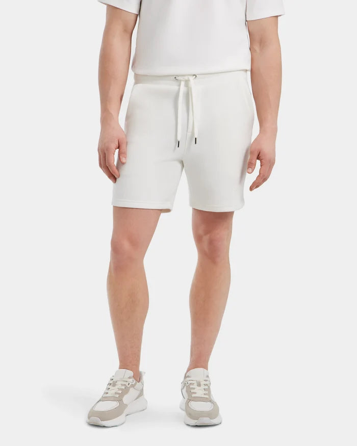 A person wears Rudsaks PASCAL - 6524022 ICELAND: an all-white, short-sleeve shirt and mens double jersey shorts with a stretch waistband, plus white sneakers with beige accents, standing against a plain white backdrop.