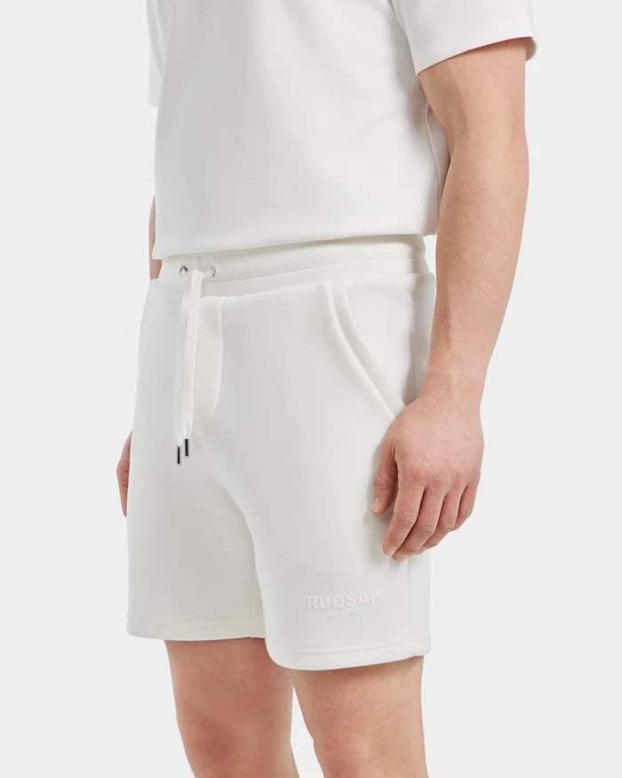 A person wears white PASCAL - 6524022 ICELAND Rudsak jersey shorts with a stretch comfort waistband and Rudsak embroidered near the hem, paired with a white short-sleeve shirt, standing with one hand by their side.