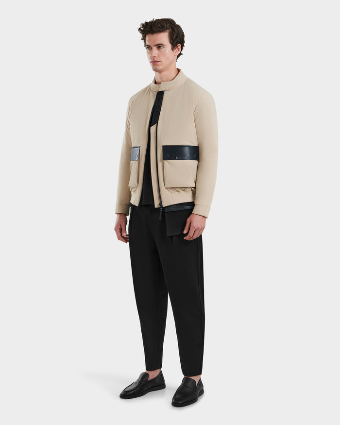 A man stands against a plain light background wearing the Rudsak PETER - 6125001 Almond beige lightweight bomber jacket with vegan leather accents, paired with a black shirt, loose black pants, and black loafers.