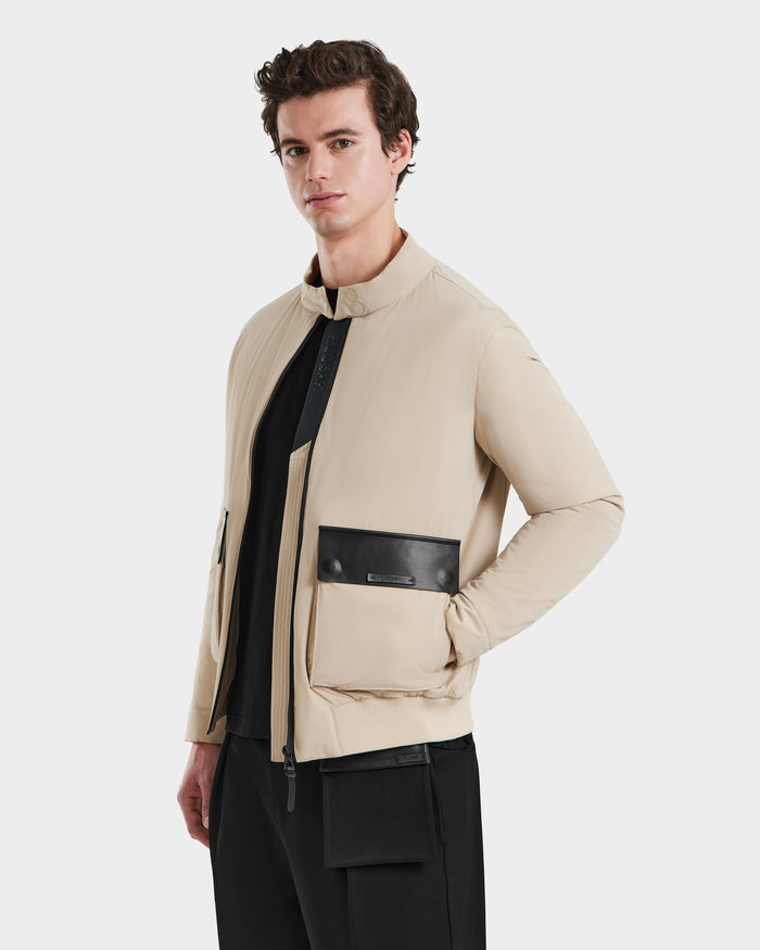 A man models the Rudsak PETER - 6125001 Almond jacket, a beige lightweight bomber with vegan leather accents, paired with black pants, as he stands against a plain white background gazing slightly to the side.