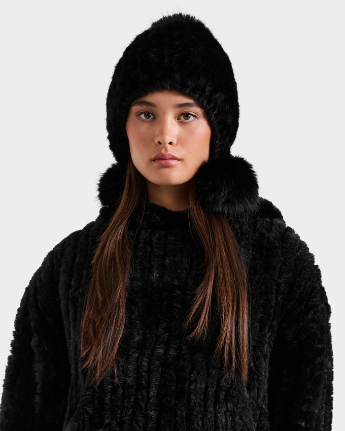 A young woman with long brown hair models the Rudsak PIA - 8425547 Black faux fur hat with pom-pom details and a matching coat, creating a chic winter look against a plain light gray background.