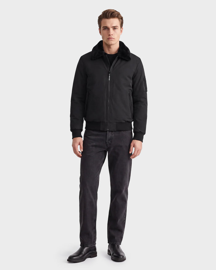 A man stands facing forward against a plain white background, wearing the Rudsak REECE - 6124592 Black men's bomber jacket with a water-resistant finish and shearling collar, paired with a black shirt, dark jeans, and black boots.