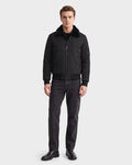 A man stands facing forward against a plain white background, wearing the Rudsak REECE - 6124592 Black men's bomber jacket with a water-resistant finish and shearling collar, paired with a black shirt, dark jeans, and black boots.