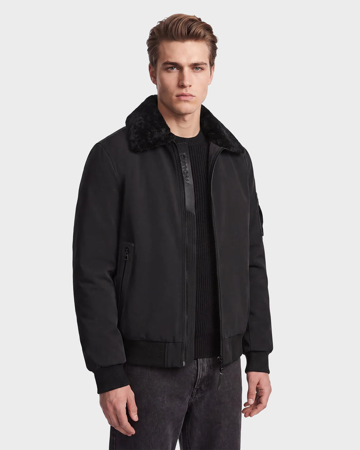 A man stands facing forward with a neutral expression, wearing the Rudsak REECE - 6124592 Black men's bomber jacket featuring a shearling collar. He pairs it with a black sweater and dark jeans against a plain light background.