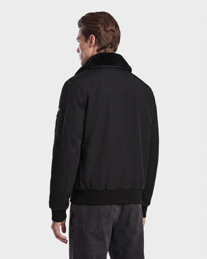 A man is standing with his back to the camera, wearing the Rudsak REECE - 6124592 Black men’s bomber jacket with a shearling collar and black jeans against a plain light background.
