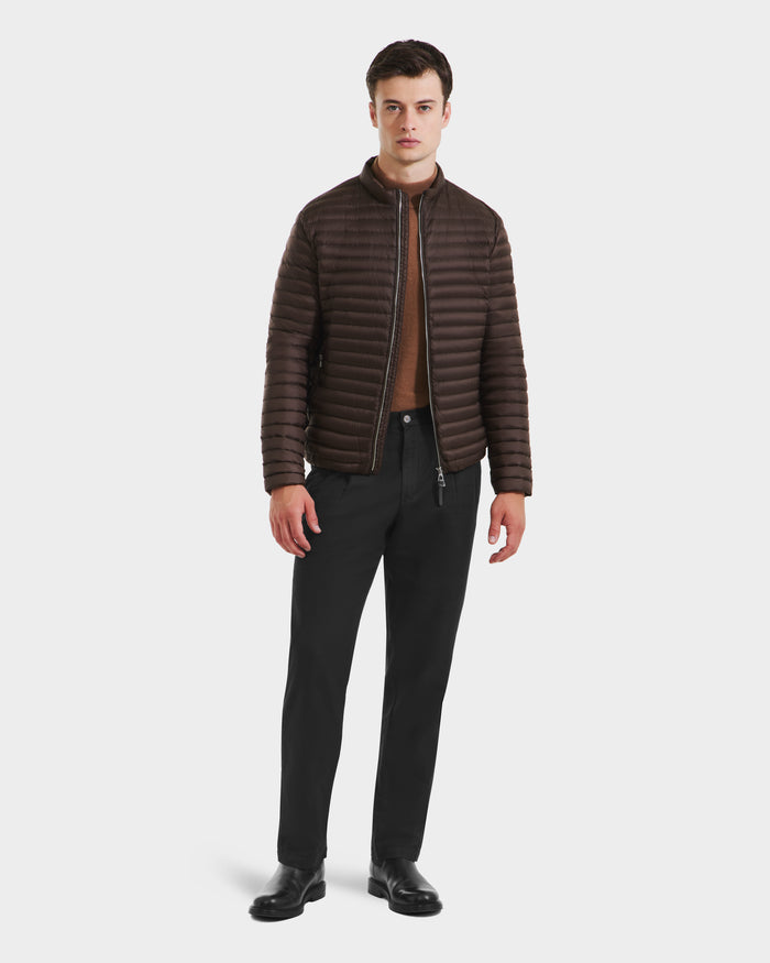 A man faces forward with a neutral expression, wearing the Rudsak REYMOND M - M125523 Mocha men’s down jacket over a brown shirt, black pants, and black shoes against a plain background. The jacket is made from recycled nylon.