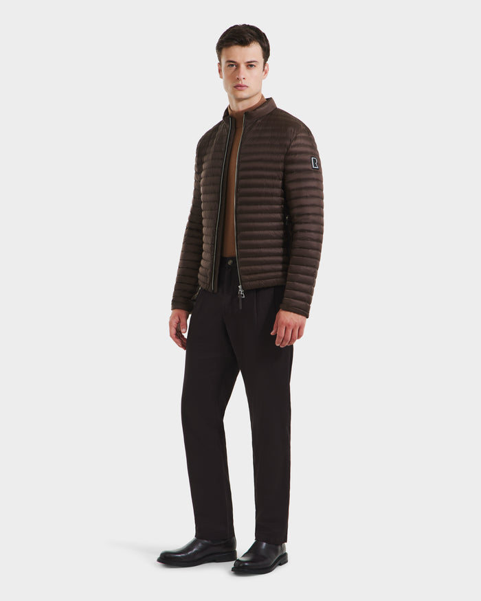 A man, facing slightly left, wears the Rudsak REYMOND M - M125523 Mocha down jacket, made from RDS-certified down and recycled nylon, over a brown shirt with dark trousers and black shoes, set against a plain white background.