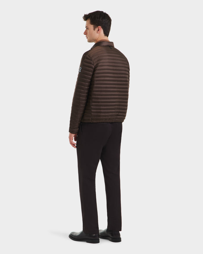 A man faces away, wearing the Rudsak REYMOND M - M125523 Mocha men’s down jacket, made from recycled nylon and RDS-certified down, paired with black pants and shoes against a plain white background.