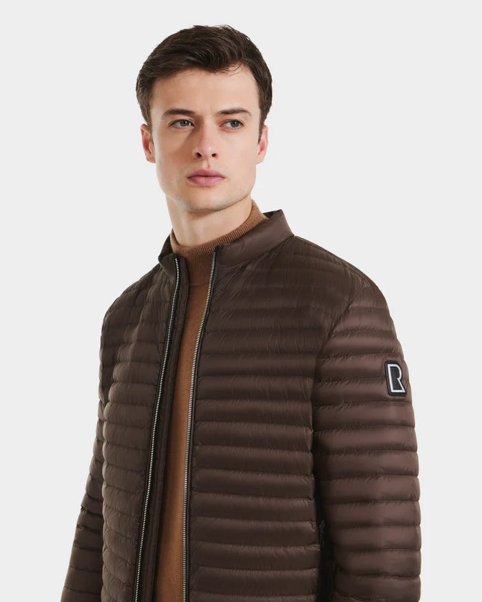 A young man with short dark hair wears the Rudsak REYMOND M - M125523 Mocha men’s down jacket made from recycled nylon over a tan sweater. He faces slightly to the side and looks away, showing the R patch on the upper sleeve.