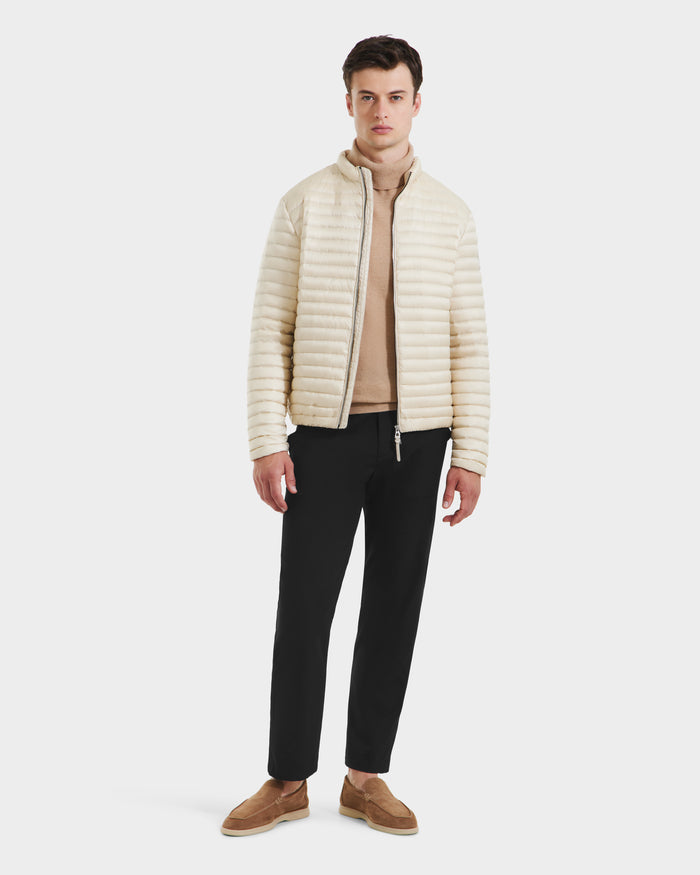 A man stands against a plain white background wearing the Rudsak REYMOND M - M125523 Vanilla men’s down jacket, filled with RDS-certified down, over a tan turtleneck, black trousers, and light brown slip-on shoes.