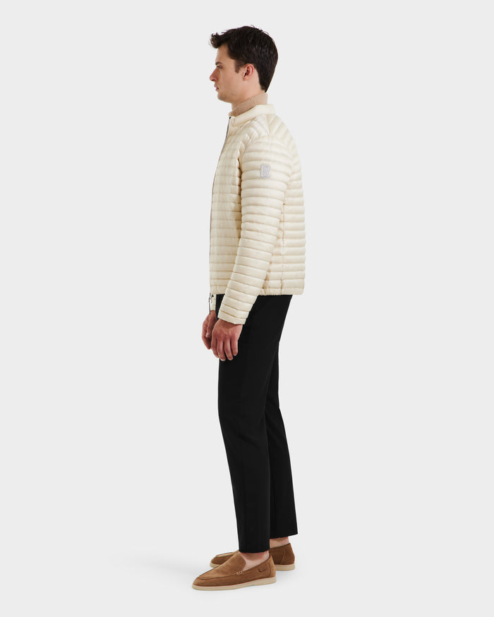 A man stands in profile against a plain background, wearing the Rudsak REYMOND M - M125523 Vanilla men’s down jacket, crafted from recycled nylon, paired with black pants and tan loafers.