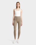 A woman faces forward in a white tank top, Rudsak’s RIANA - 8524031 Cement taupe stretch leggings, and white sneakers against a light gray background—the ideal athleisure look.