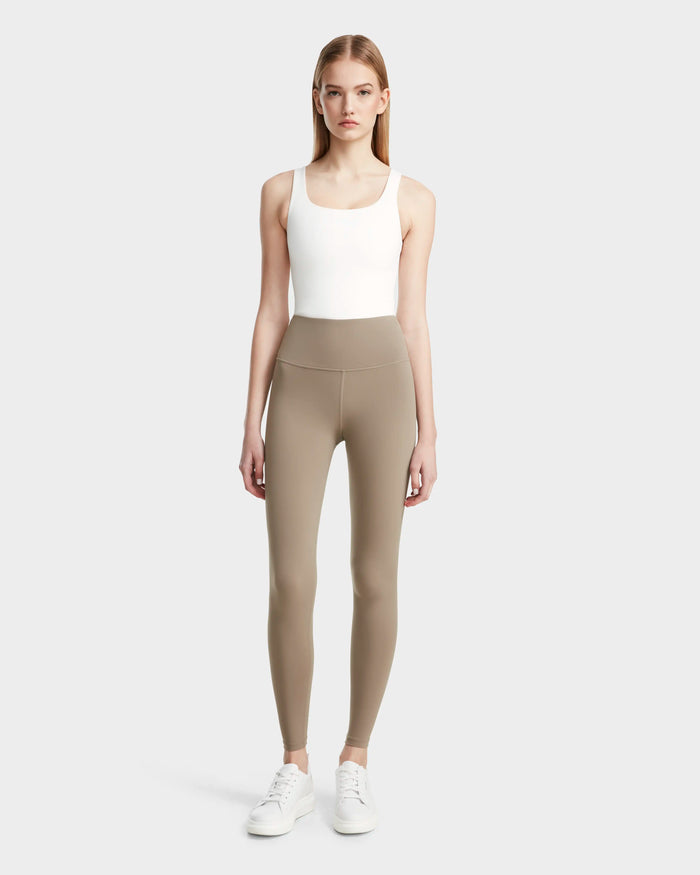 A woman faces forward in a white tank top, Rudsak’s RIANA - 8524031 Cement taupe stretch leggings, and white sneakers against a light gray background—the ideal athleisure look.