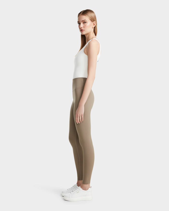 A woman stands in profile against a plain background, wearing Rudsak's RIANA - 8524031 Cement high-waisted women's stretch leggings with a white sleeveless top and sneakers. Her straight, light brown hair frames her neutral expression.