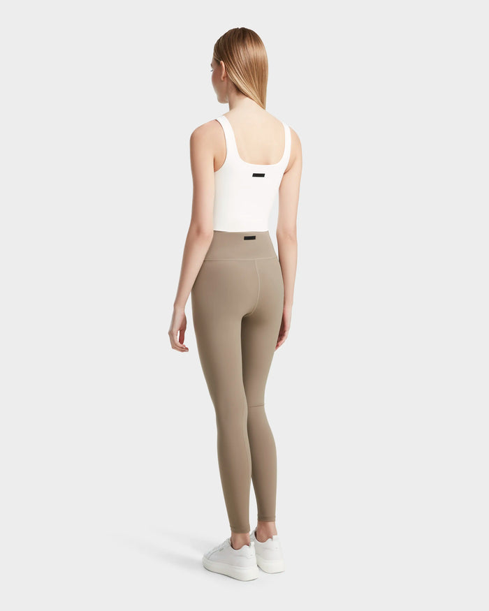 A woman with straight blonde hair faces away, wearing the RIANA - 8524031 Cement high-waisted stretch leggings by Rudsak and a white tank top—her athleisure style completed against a light gray background.