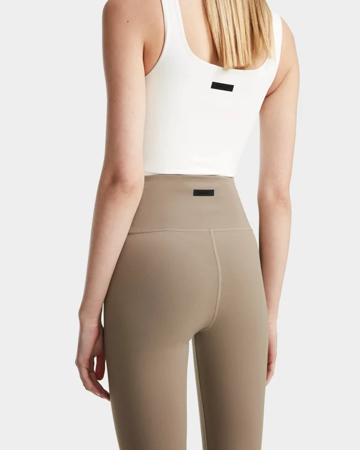 A person with straight blonde hair is seen from behind, wearing a white sleeveless top and high-waisted Rudsak RIANA - 8524031 Cement women's stretch leggings, both featuring small black rectangular labels on the back.
