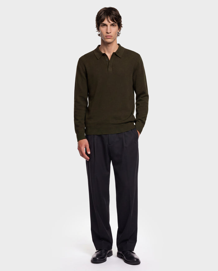 A person stands facing forward against a plain white background, wearing the Rudsak RICHARD - 6524529 Olive men’s knit polo sweater with long sleeves, paired with black pleated trousers and black shoes.