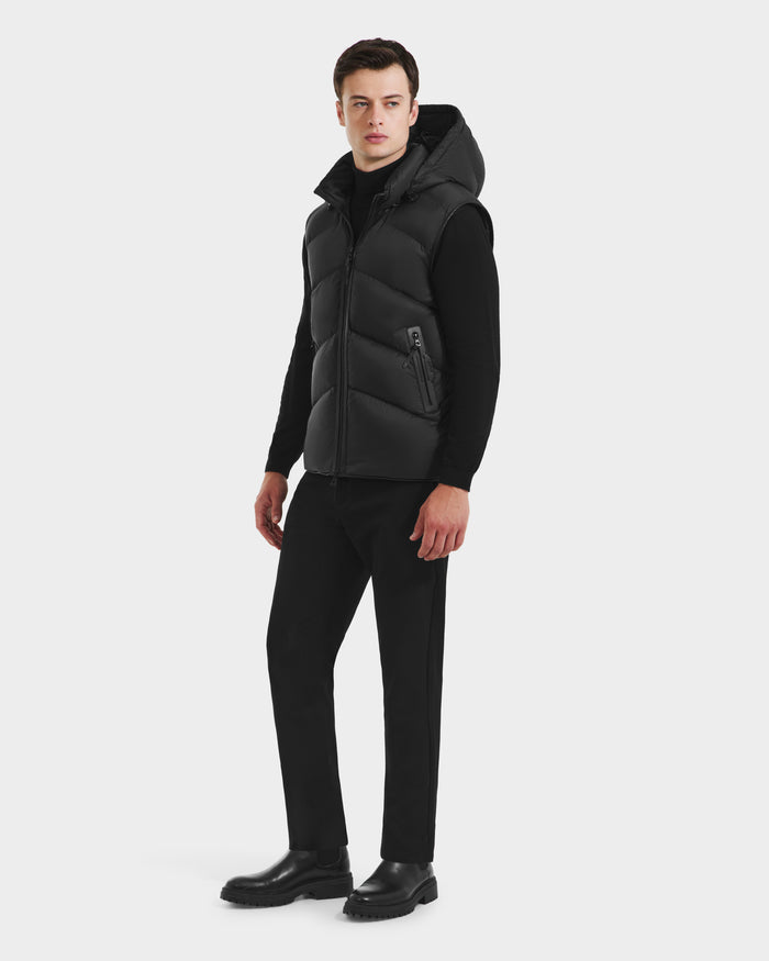 A man in the Rudsak RICHARDSON - M125527 Black men’s hooded vest stands against a plain light background, facing slightly to the side, styled with a black turtleneck, black pants, and black boots.