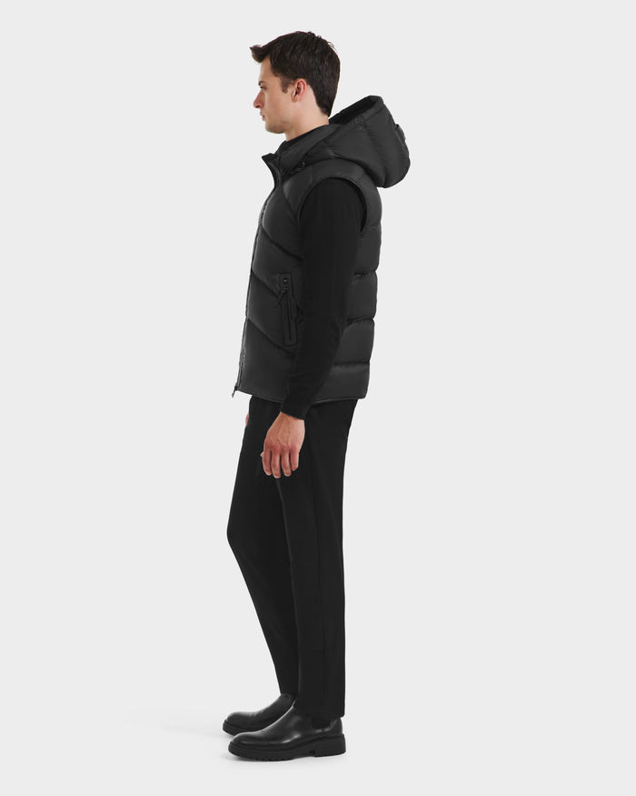 A man stands in profile wearing the Rudsak RICHARDSON - M125527 Black men’s hooded vest for lightweight warmth and water-repellent comfort, layered over a black long-sleeve shirt, black pants, and boots, against a plain white background.