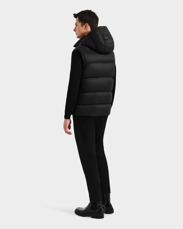 A man faces away, wearing the Rudsak RICHARDSON - M125527 Black men’s hooded puffer vest over a black long-sleeved shirt, black pants, and boots against a plain light gray background.