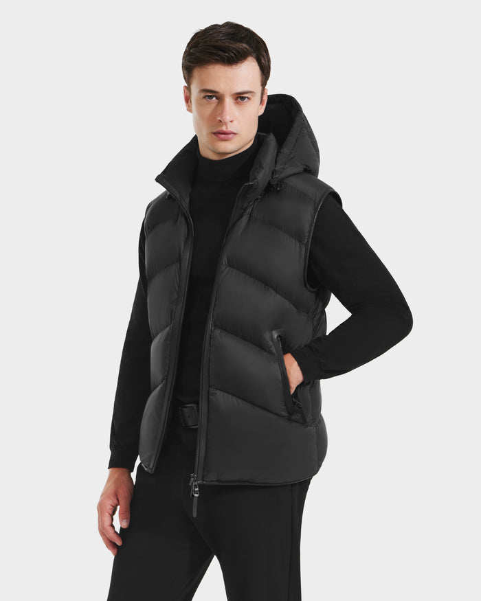 A man models the Rudsak RICHARDSON - M125527 Black men’s hooded vest over a black turtleneck and pants, one hand in his pocket, highlighting its lightweight warmth and water-repellent style against a light background.
