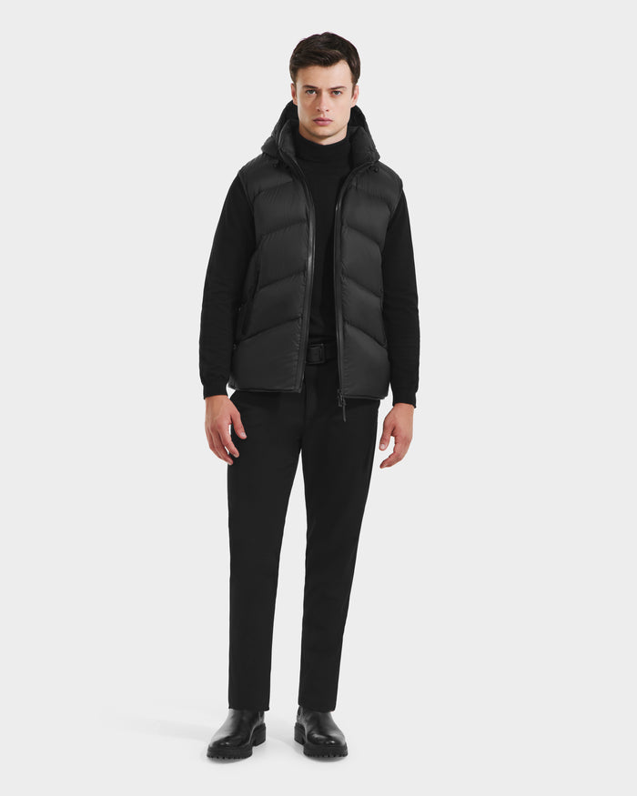 A man faces forward against a white background, wearing the Rudsak RICHARDSON - M125527 Black men’s hooded vest over a black long-sleeve shirt, black pants, and black shoes for lightweight warmth.