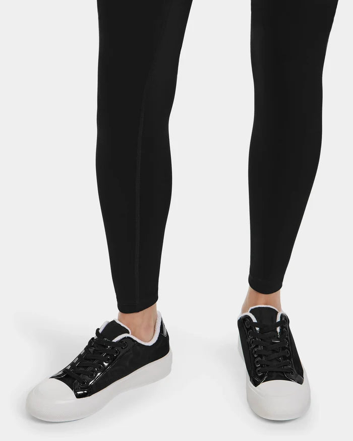 A person wearing black leggings and Rudsak RIMA - 8225001 Black Pattern womens platform sneakers with black patent leather uppers and white soles, standing on a white background.