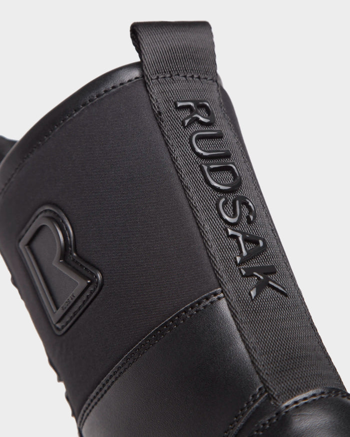 Close-up of the Rudsak RITA - L222502 Black womens winter boot with weather-treated leather, a raised RUDSAK logo on the pull tab, and an embossed R emblem on the upper, shown against a plain white background.