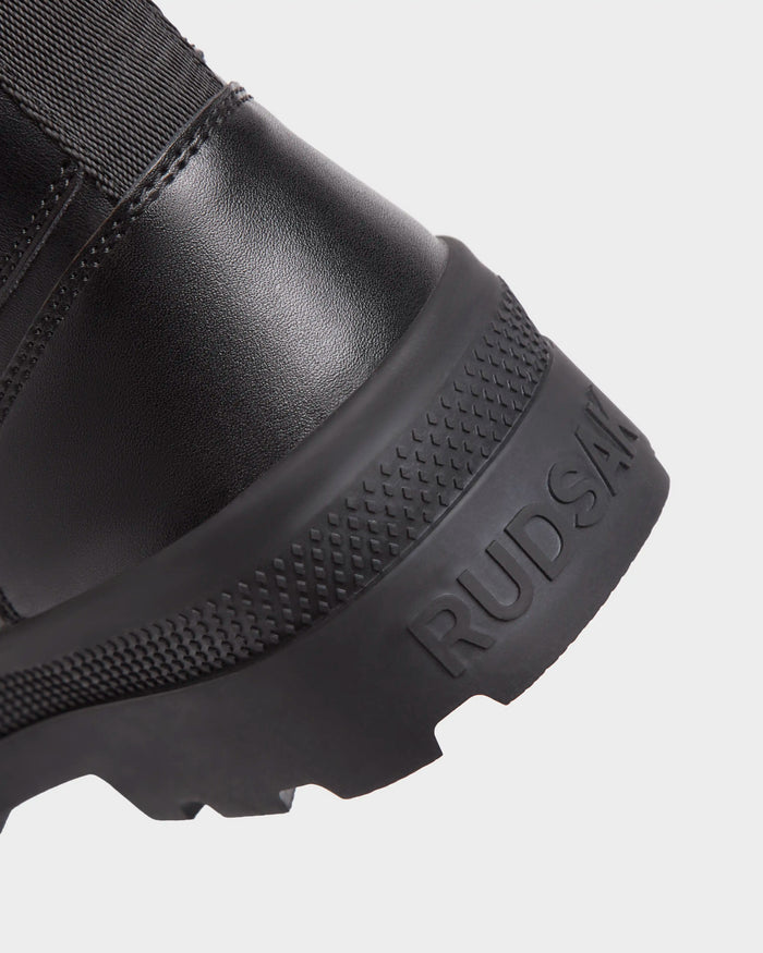 Close-up of the heel and sole of the black RITA - L222502 womens winter boot by Rudsak, featuring textured rubber, weather-treated leather, and the Rudsak name embossed on the side of the sole.