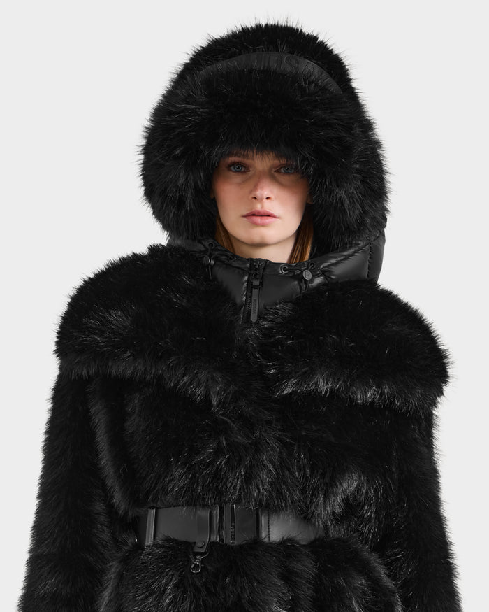 A person wears the Rudsak ROMINA - 8425545 Black coat with a glossy faux fur finish, hood, high collar, and belt detail, paired with a matching faux fur bucket hat for a stylish cozy-luxe look against a light background.