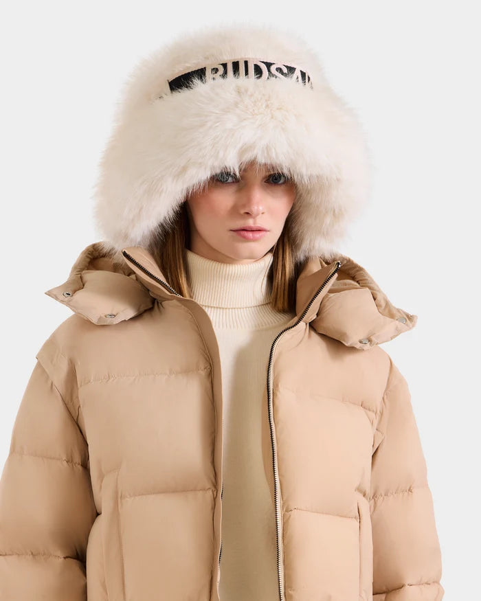 A woman wears the Rudsak ROMINA - 8425545 Vanilla puffer jacket and a white faux fur bucket hat with black lettering, standing against a plain light backdrop.
