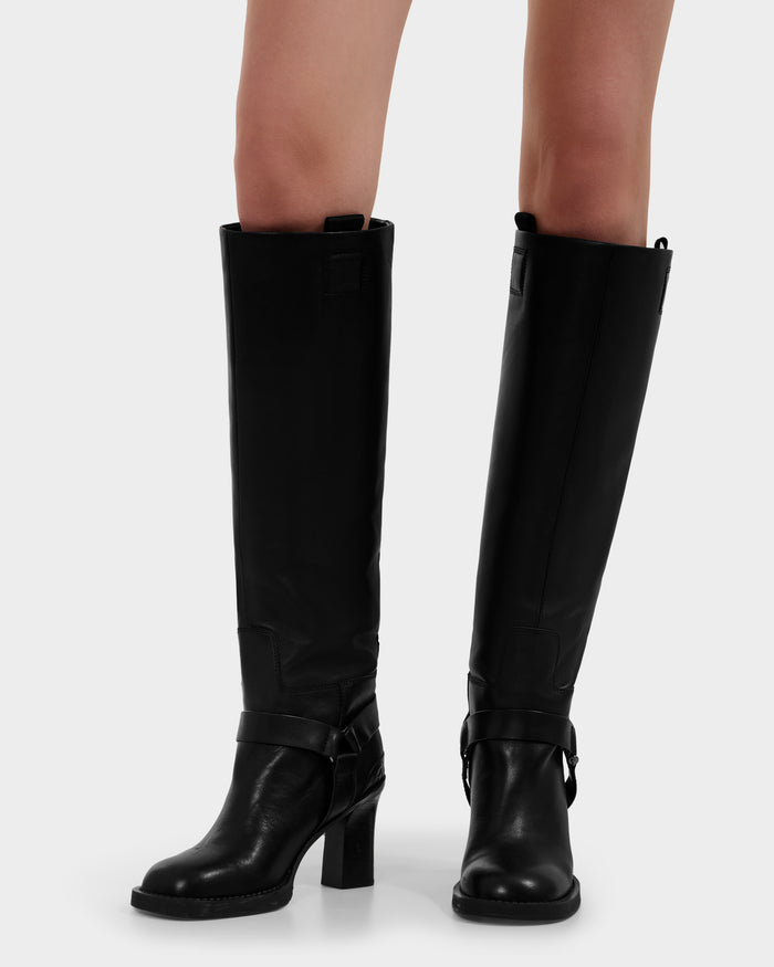 A person wearing Rudsak's ROSSEY - 8225574 Black knee-high leather heeled boots with side straps, standing on a neutral background. Only the lower legs and feet are visible.