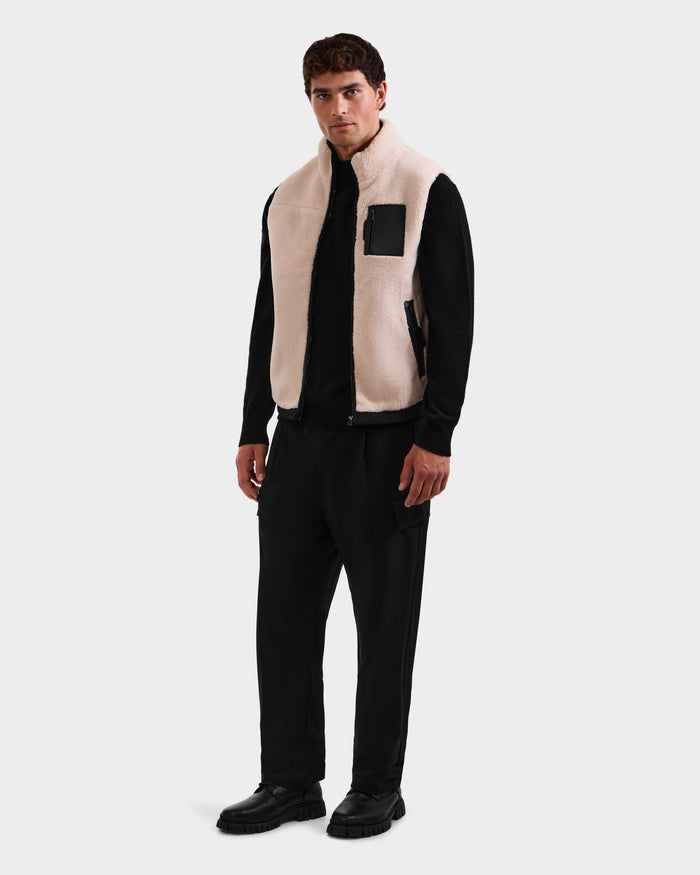 A man stands against a plain light gray background wearing the Rudsak RUMMY - U524502 Vanilla M zip-up vest with a black chest pocket, styled with a black long-sleeve shirt, black pants, and black shoes.