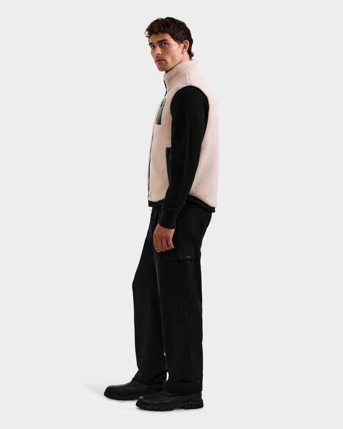 A man stands sideways against a light gray background wearing Rudsak’s RUMMY - U524502 Vanilla M unisex teddy vest over a black long-sleeve shirt, black cargo pants, and black shoes.