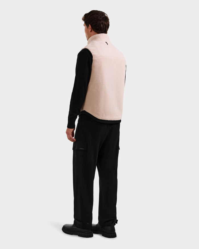 A man faces away, wearing Rudsak's RUMMY - U524502 Vanilla M unisex teddy vest over a black long-sleeve shirt and cargo pants with black shoes. The zip-up vest adds a cozy accent against the light background.