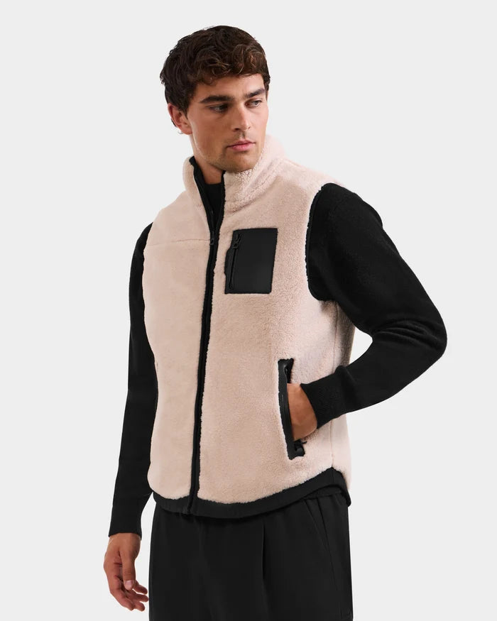 A man wearing the Rudsak RUMMY - U524502 Vanilla M unisex teddy vest with black zipper pockets over a black long-sleeve shirt and pants stands against a plain light background, looking to the side.