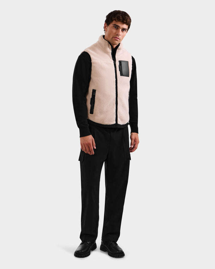 A man stands against a plain background wearing the Rudsak RUMMY - U524502 Vanilla M light beige zip-up vest with a black zipper and pocket, over a black long-sleeve shirt, pants, and shoes.
