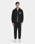 A man stands facing forward, wearing the Rudsak RYAN - 6125023 Black quilted jacket with eco-friendly insulation, paired with black pants, a white shirt, and white sneakers against a plain light background.