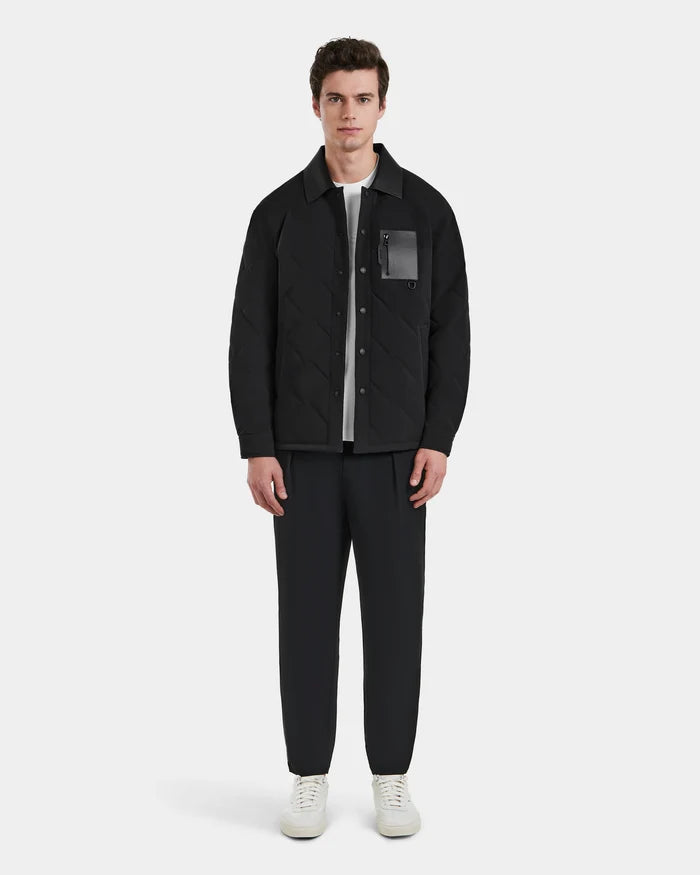 A man stands facing forward, wearing the Rudsak RYAN - 6125023 Black quilted jacket with eco-friendly insulation, paired with black pants, a white shirt, and white sneakers against a plain light background.