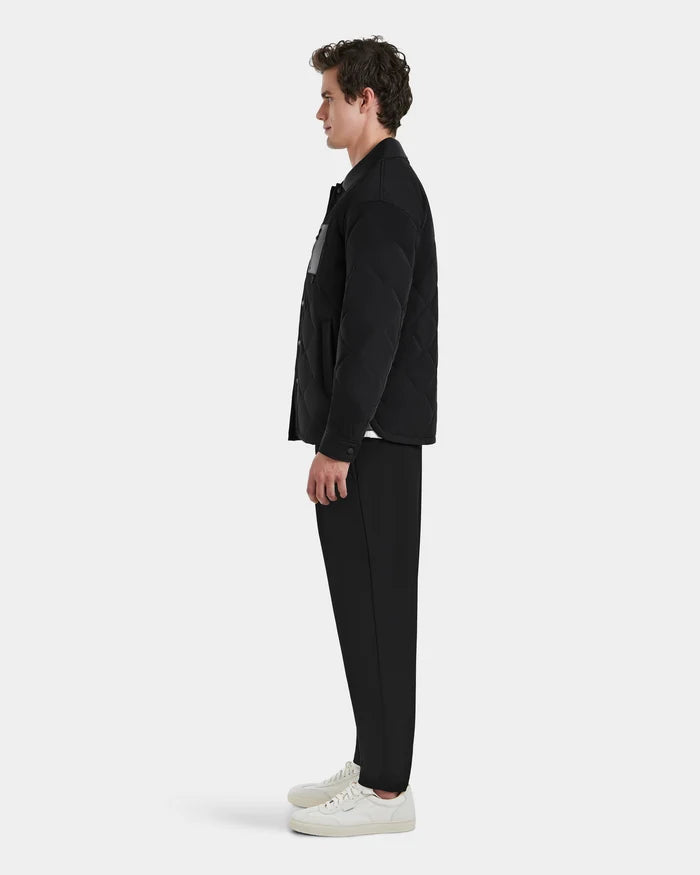 A man stands in profile against a plain background, wearing the Rudsak RYAN - 6125023 Black quilted jacket with eco-friendly insulation, paired with black pants, a white shirt, and white sneakers.