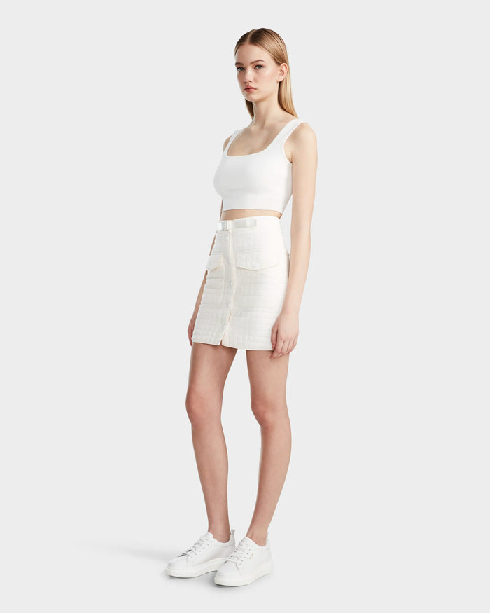 A woman with long blonde hair and a neutral expression wears the Rudsak SABINE - 8524063 Eggshell: a white crop top and quilted short skirt with buckle webbing front closure, creating a standout women’s statement look against a plain background.
