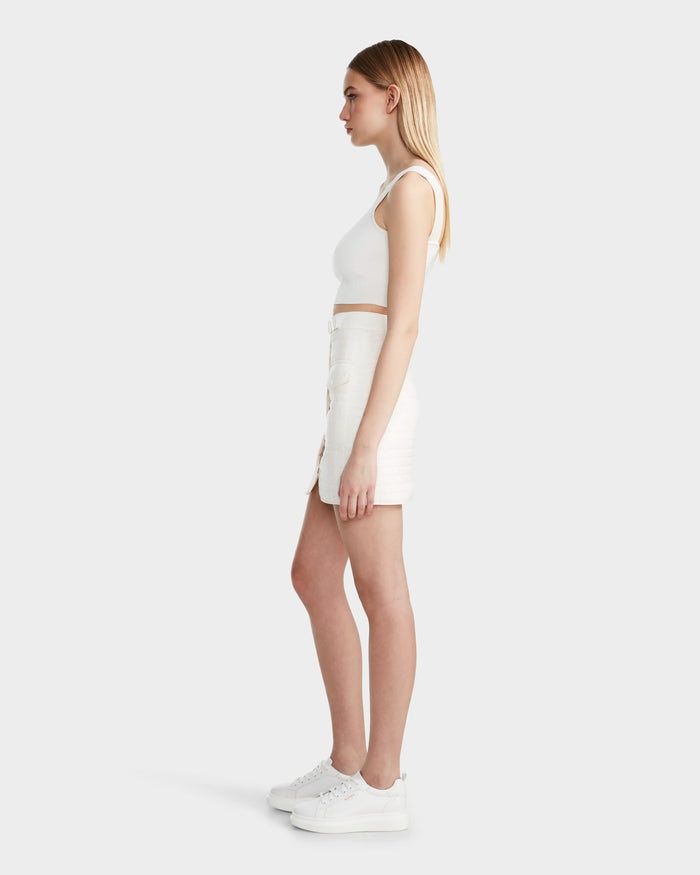A young woman with straight, blonde hair stands in profile against a plain background, wearing the Rudsak SABINE - 8524063 Eggshell sleeveless crop top and a quilted short skirt as a statement piece, paired with white sneakers.