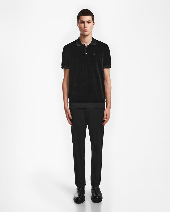 A young man stands against a white background, wearing the Rudsak SAM - 6524502 Black polo with black pants and shoes, his arms relaxed at his sides for a modern luxury mens look.