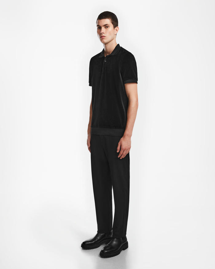 A young man stands against a plain white background, wearing the Rudsak SAM - 6524502 Black polo with matching black trousers and dress shoes. He faces slightly to the side, showing off this luxury mens fashion look with a neutral expression.