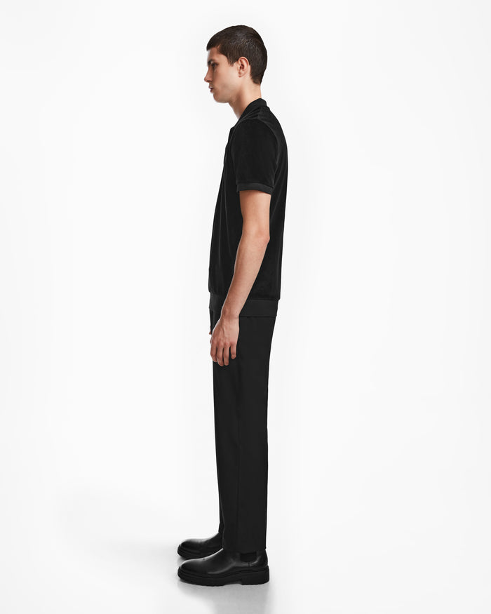 A young man stands in profile against a white background, wearing the SAM - 6524502 Black polo by Rudsak, paired with black pants and boots. He looks straight ahead with relaxed arms, embodying refined luxury mens fashion.