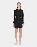 A woman stands facing forward in the Rudsak SETA - 8525019 Black, a contemporary black long-sleeve mini dress with white striped sleeves, a black belt at the waist, and white sneakers, styled against a plain white background.