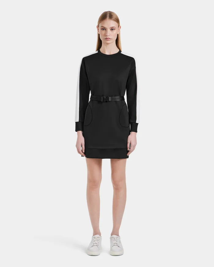 A woman stands facing forward in the Rudsak SETA - 8525019 Black, a contemporary black long-sleeve mini dress with white striped sleeves, a black belt at the waist, and white sneakers, styled against a plain white background.