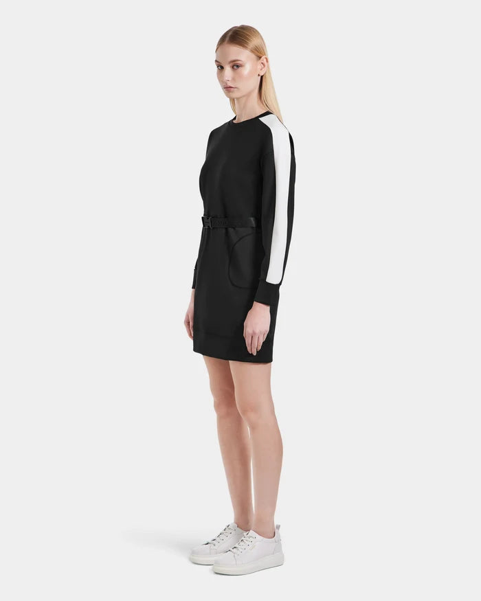 A woman models the Rudsak SETA - 8525019 Black, a women’s relaxed-fit black long-sleeve dress with white panel sleeves and a belted waist, styled with white sneakers. She poses against a plain white background.