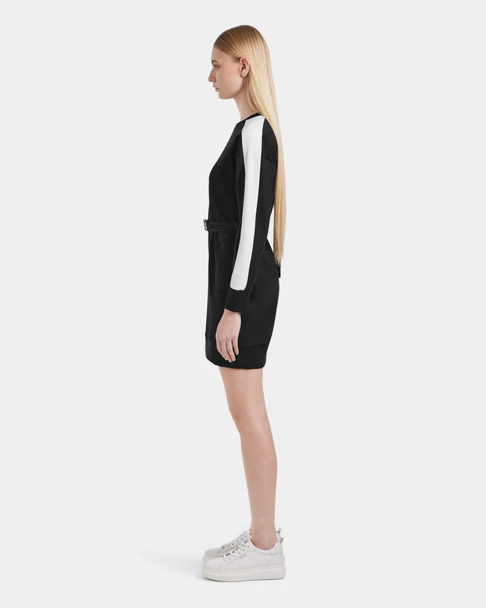 A woman with long straight blonde hair stands in profile, wearing the Rudsak SETA - 8525019 Black mini dress with white sleeve stripes and white platform sneakers, set against a plain white background.