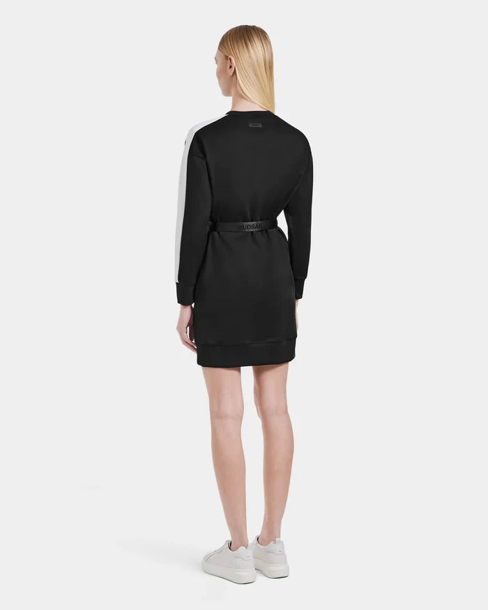 A woman, with straight blonde hair past her shoulders, faces away wearing Rudsak’s SETA - 8525019 Black women’s relaxed-fit dress with a white sleeve stripe and cinched waist, paired with white sneakers against a plain background.